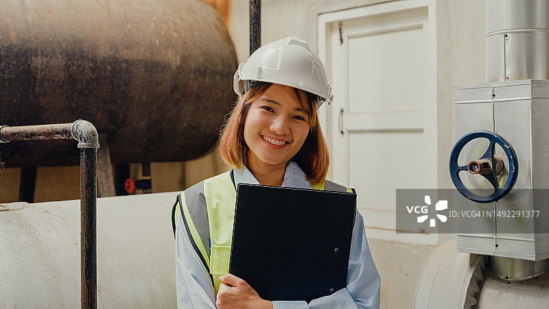 年轻的亚洲女工业工程师戴着安全帽，在化工厂的蒸汽锅炉附近对着镜头微笑。重工业和工程师。图片素材