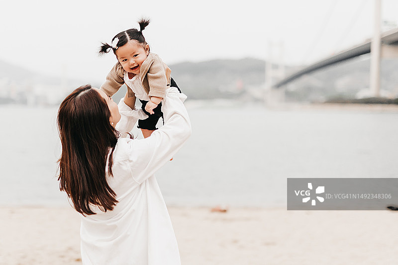 亚洲母亲和她的幼儿在海滩上度过美好的一天。幼儿在沙滩上玩得很开心，母亲面带微笑地看着图片素材