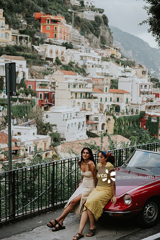 两个年轻漂亮的女人坐在红色阿尔法罗密欧老爷车的引擎盖上，背景是波西塔诺的景色图片素材