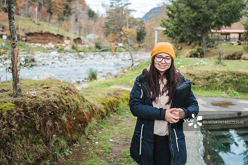 穿着保暖的年轻数字游民女性，带着笔记本电脑在温泉区行走图片素材