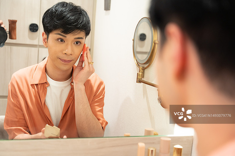 亚裔男子身穿橙色服装，坐在镜子前，拿着粉扑给自己化妆图片素材