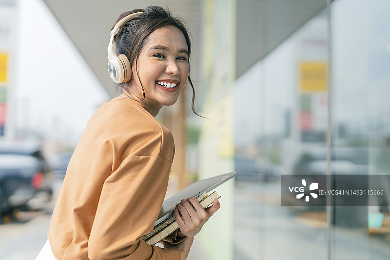 微笑的亚洲少女愉快地打招呼，手势友好地向镜头挥手，同时在户外商店店面白天听着耳机里的音乐图片素材