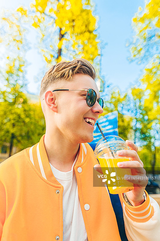开心的年轻男人穿着黄色夹克戴墨镜漫步城市享受鲜榨橙汁图片素材