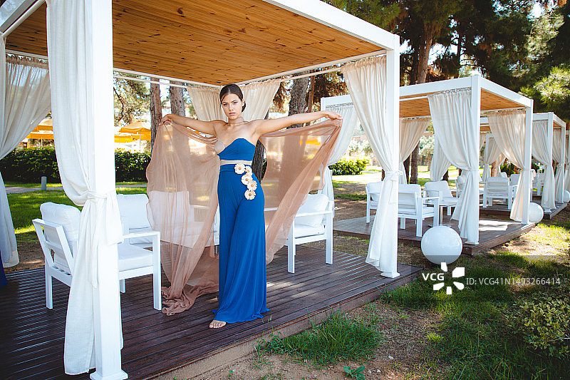 年轻时尚模特身穿蓝色无肩带长裙，站在公园凉亭旁图片素材