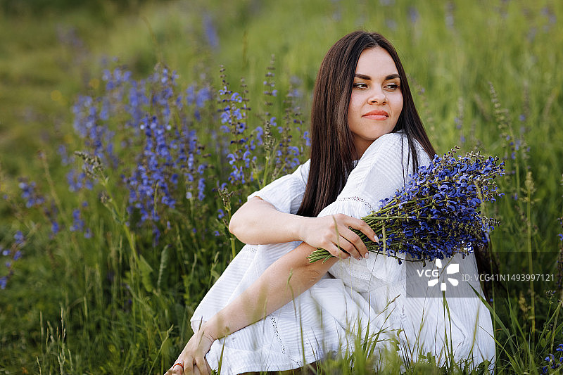 年轻美丽的棕发女子穿着白色连衣裙坐在春天开满羽扇豆的绿色草地上图片素材