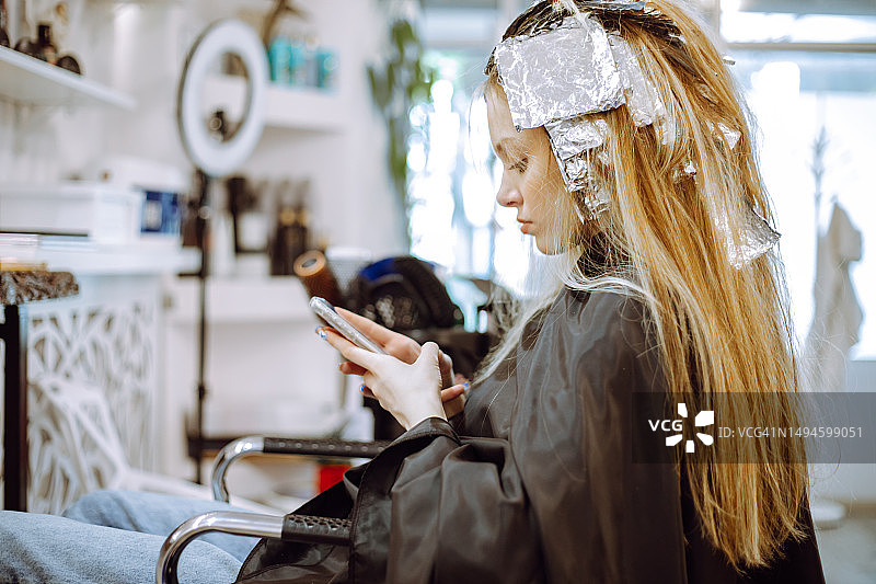 在美发店染发时，坐着使用手机的少女侧视图图片素材