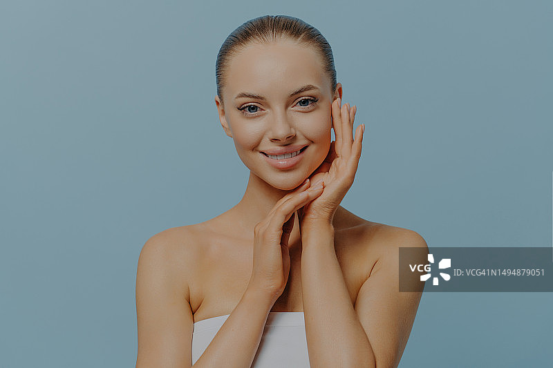 触碰脸颊，肌肤柔嫩健康的年轻漂亮女人，半裸站在蓝色背景前，英国图片素材