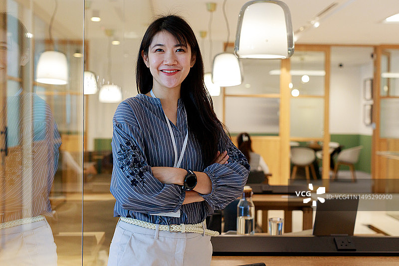 一位自信的年轻亚洲女性的肖像，她面带灿烂的笑容，双臂交叉地靠在办公桌旁，散发着积极的气息图片素材