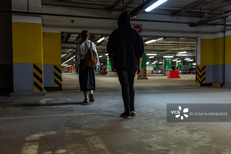身穿连帽衫的年轻男人走在空旷的地下停车库里，身后是一名年轻女子图片素材