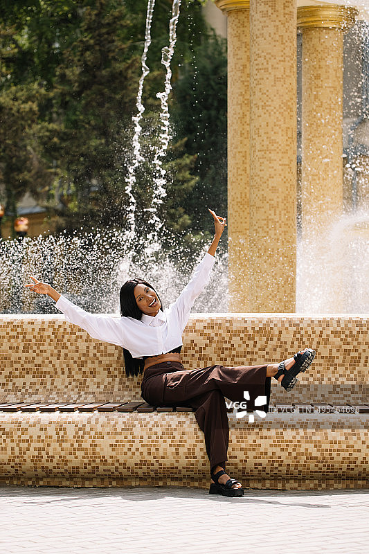 坐在喷泉附近，举起双臂微笑的快乐的多种族黑发女人图片素材