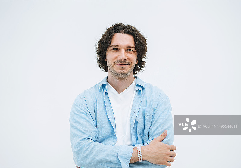 微笑的年轻深色头发男子肖像，身穿蓝色衬衫，在白色背景下看着相机图片素材
