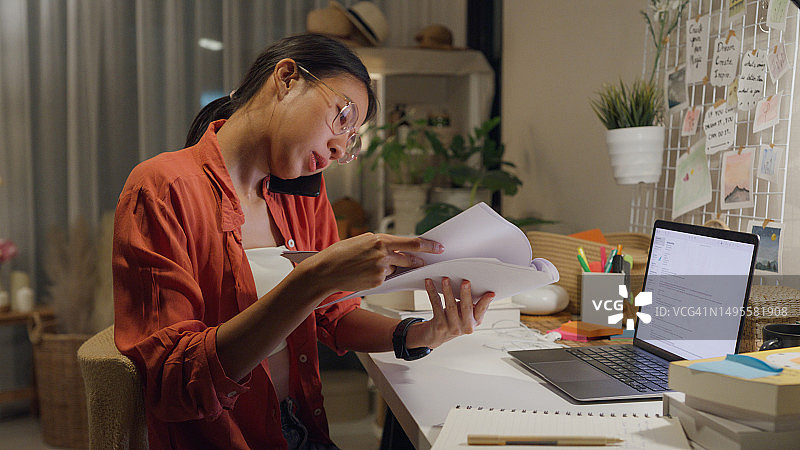 年轻的亚洲女性穿着休闲装坐在桌子前，用笔记本电脑和手机在客厅里与同事开会。在家工作。图片素材