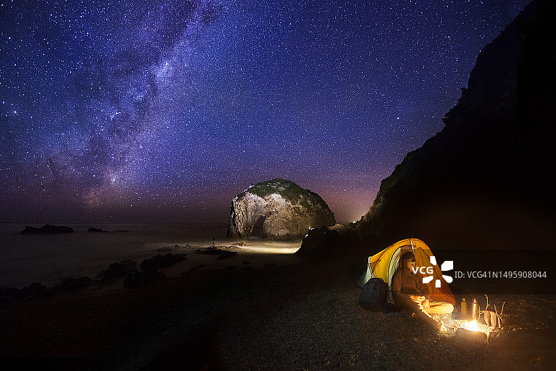 男子在银河系下，篝火旁露营，独特的岩石构成一匹马在背景中饮水图片素材