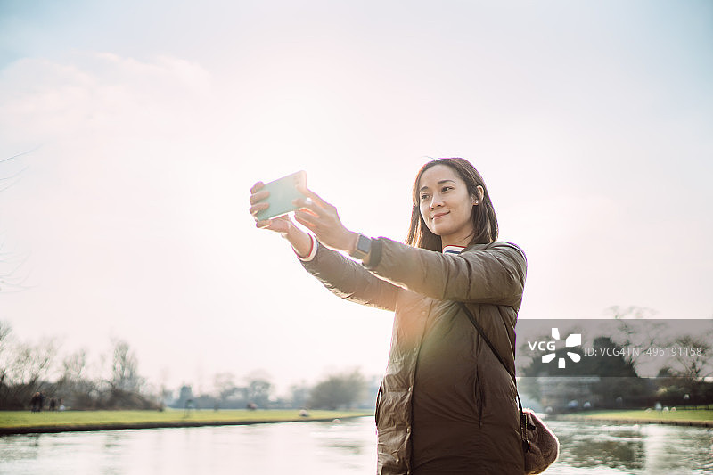 年轻女游客在公园湖边用智能手机自拍图片素材