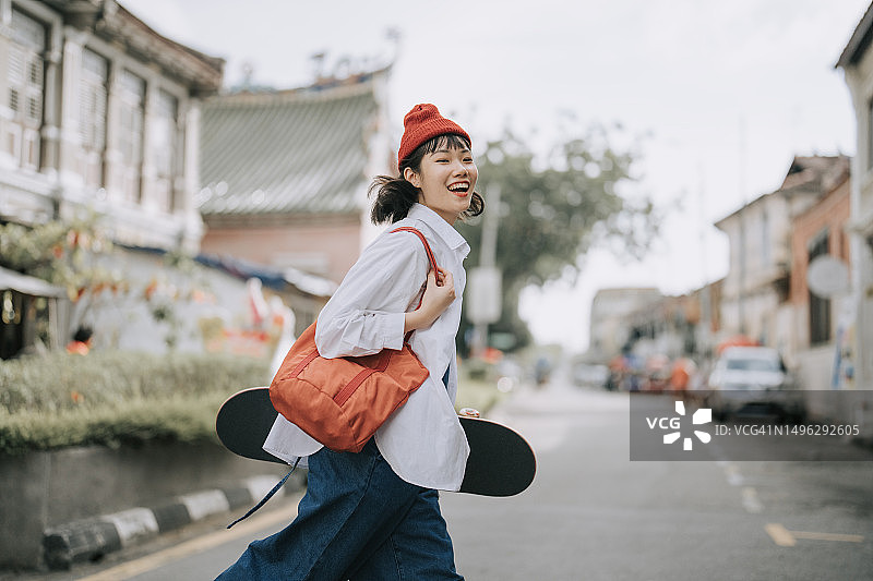 快乐的中国年轻女子在老城区过马路，手拿滑板图片素材
