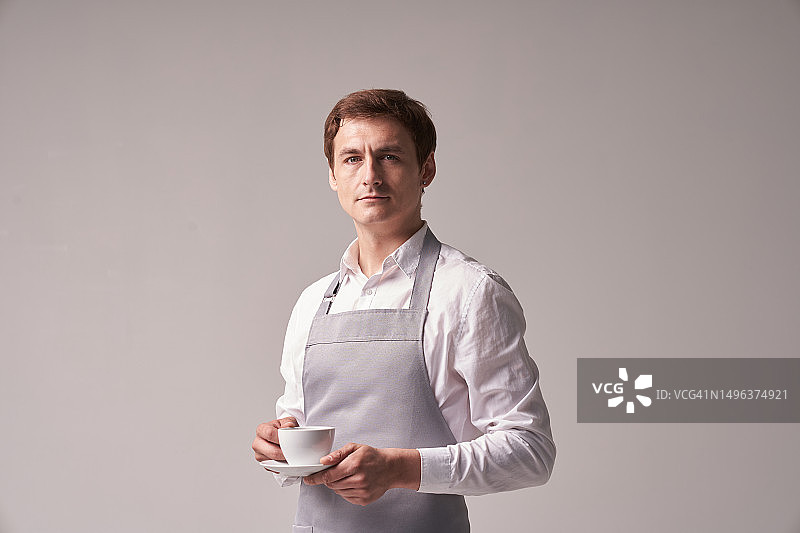 灰色背景下积极的男服务员，咖啡师为顾客端咖啡，成功的年轻人肖像图片素材