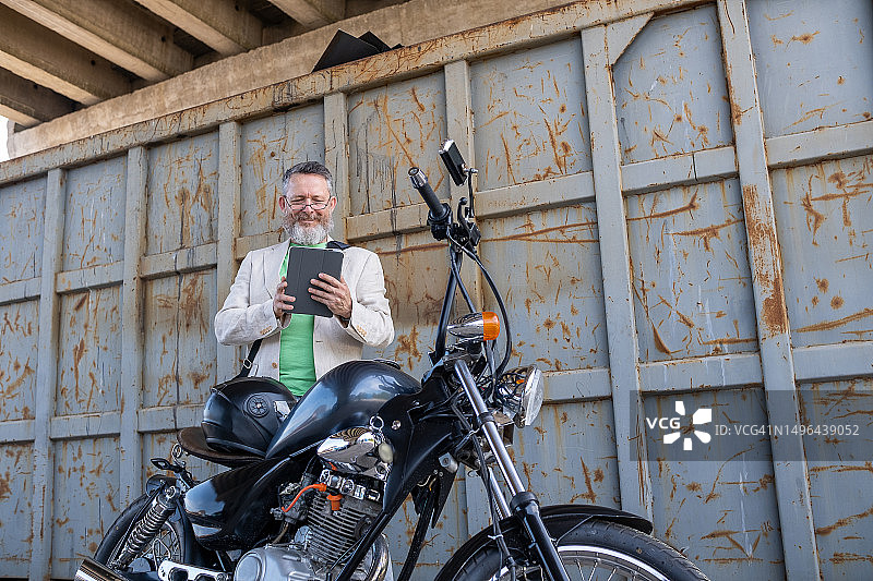 中年男人身穿浅绿色T恤、米色夹克和灰色牛仔裤，站在Bobber摩托车后图片素材