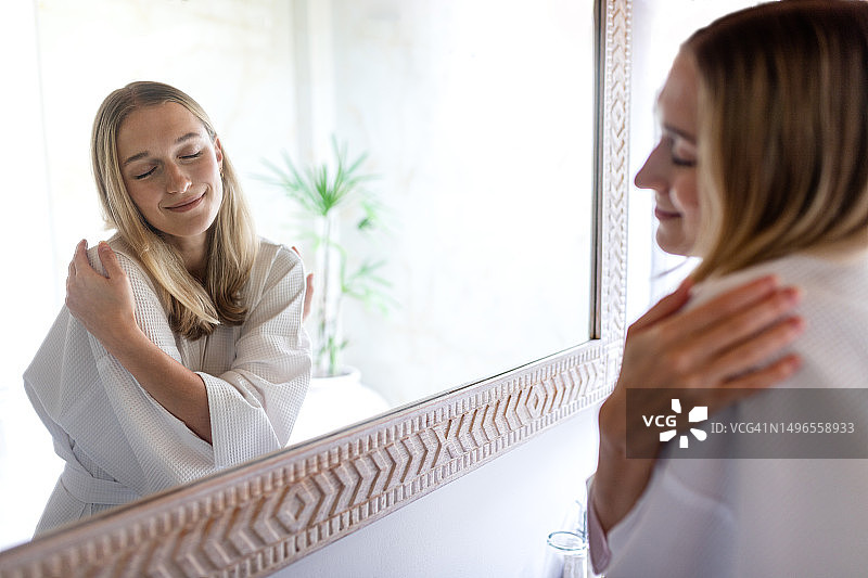 闭着眼睛在镜子前拥抱自己的年轻金发女子。女性表达自爱，穿着浴袍，在浴室镜中拥抱自己，感到放松，充满自信图片素材