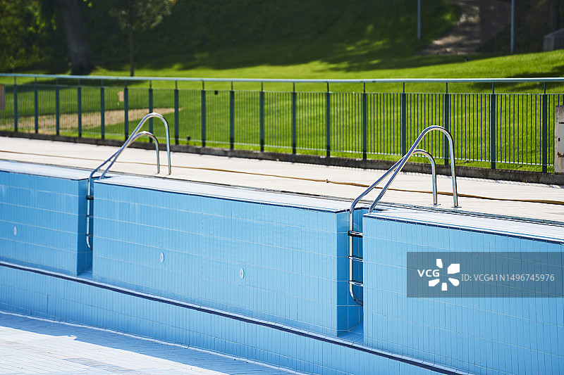 公园里没有水的蓝色空游泳池特写，带绿色草地和金属楼梯，正面视角图片素材