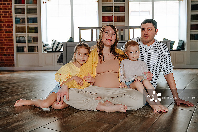 一家人坐在地板上。女人怀孕了。他们看着镜头。图片素材