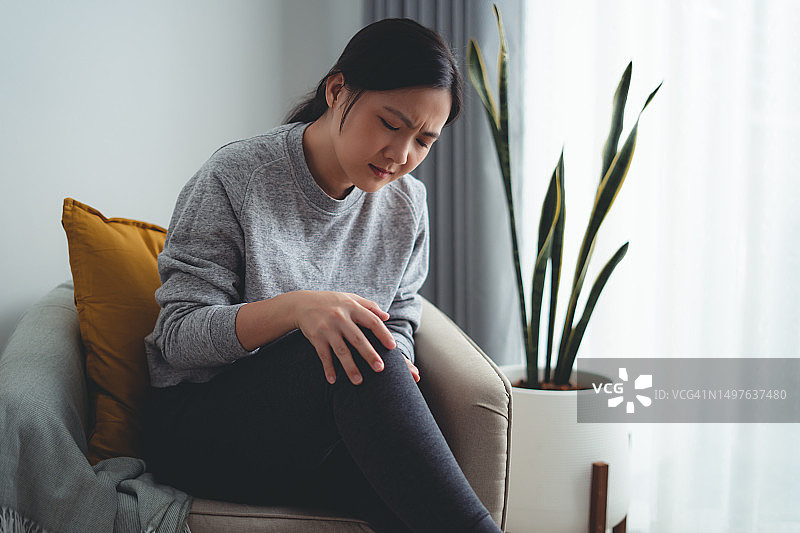 亚洲妇女坐在扶手椅上，在家中客厅感到膝盖疼痛图片素材