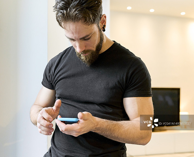 留着胡须的成年男子站在现代公寓里，专心致志地在手机上查看社交网络。图片素材