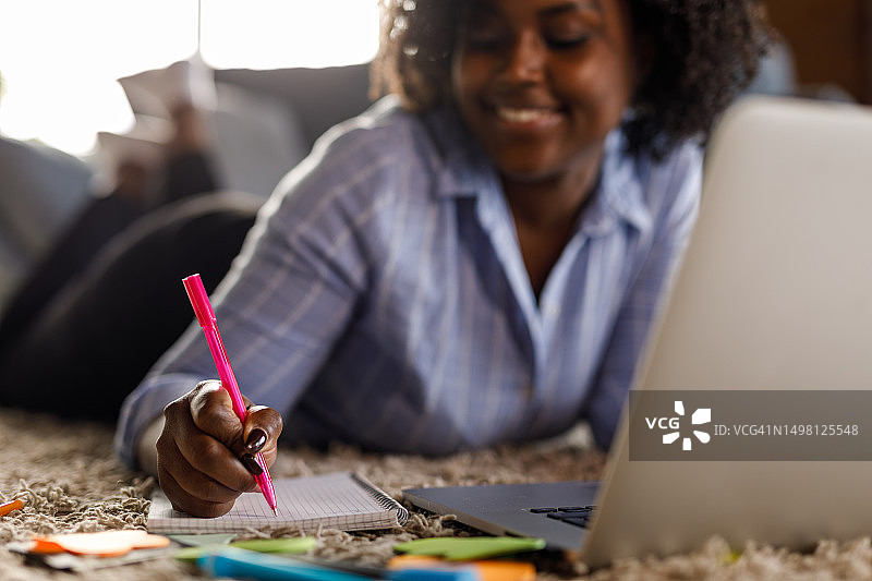 年轻女子趴着使用笔记本电脑并做笔记，为大学做研究图片素材