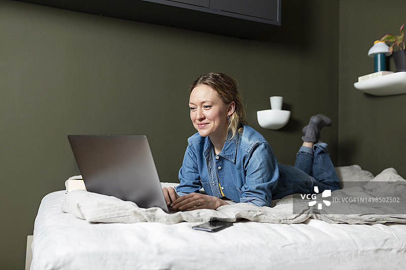 自由职业者穿着牛仔衬衫在家里的床上用笔记本电脑工作图片素材