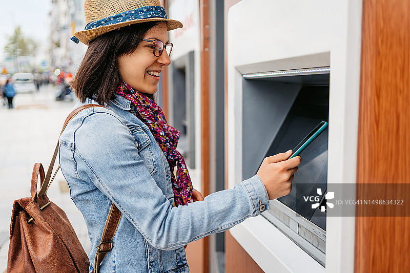 在伊斯坦布尔的自动取款机上用手机提取土耳其里拉的年轻女人图片素材