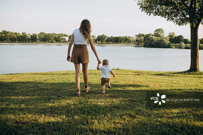 母亲牵着一岁孩子的手沿着草地走向湖边，背面视角图片素材