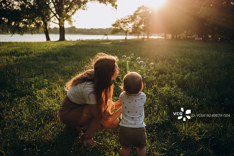 夕阳下，卷发母亲和孩子在湖岸边吹肥皂泡。背影图片素材