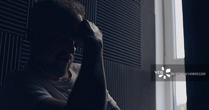 沮丧的男子独自一人在黑暗的房间里，坐在窗边揉着鼻梁图片素材