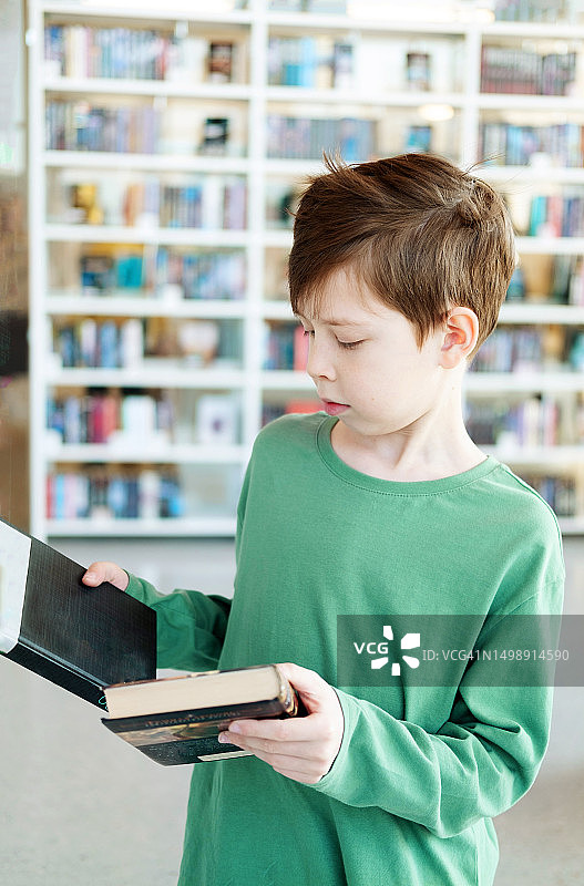 一个穿着绿色T恤的蓬松男孩站在图书馆里，为自己选择一本书图片素材