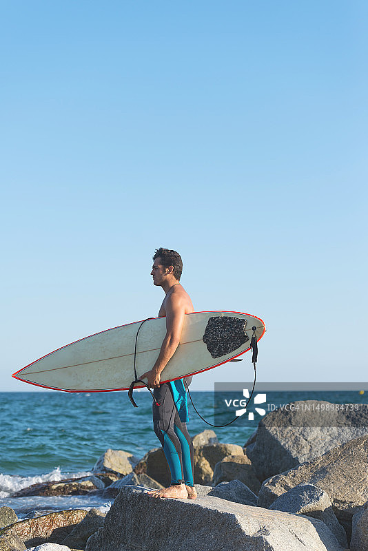 冲浪男子站在海中岩石上，手持冲浪板图片素材