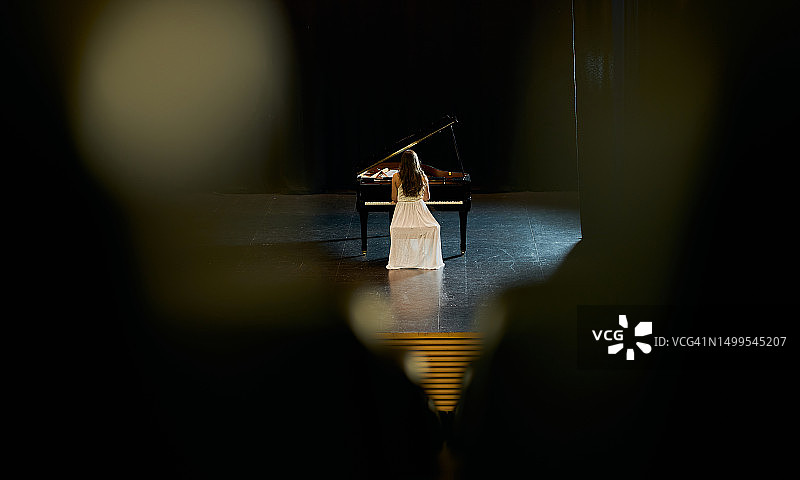 身穿白色连衣裙的女孩在舞台上背对着观众演奏黑色三角钢琴图片素材