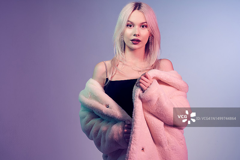 一个蓝色和紫色背景下的年轻金发女郎。一位身穿黑色连衣裙和白色毛皮大衣的女人，化着精致的妆容，发型时尚。工作室里的时尚美女故事。图片素材
