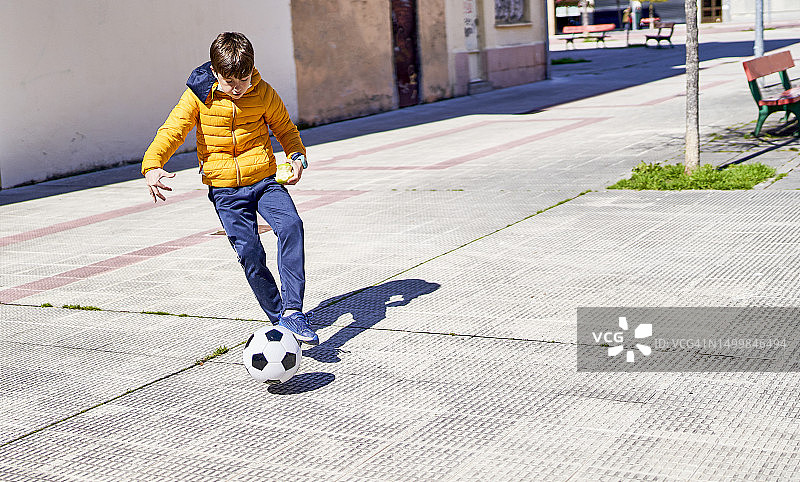 阳光明媚的一天，一个男孩穿着外套在自家车库里踢足球的正面照图片素材