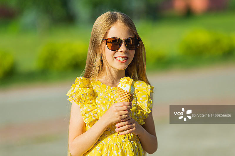 戴着太阳镜、身穿黄色连衣裙的金发女孩吃冰淇淋。夏季生活方式，童年图片素材