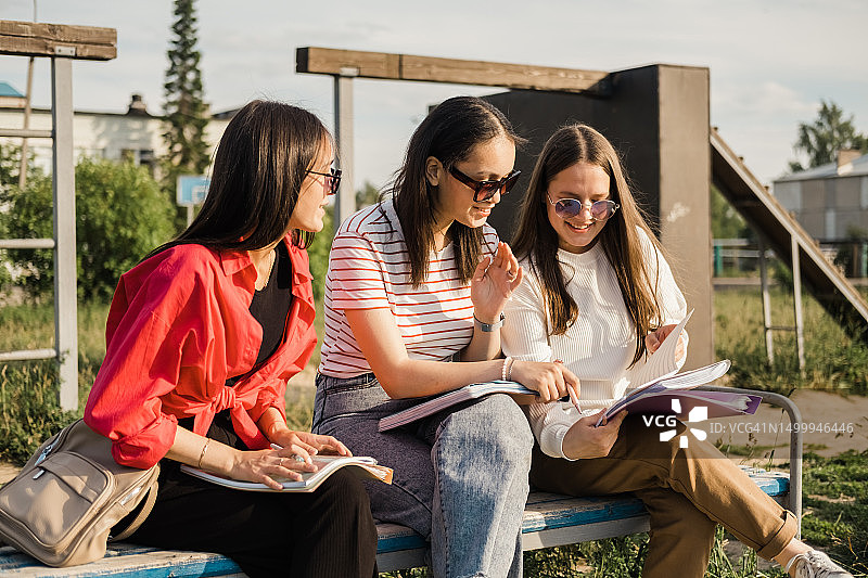 在一个阳光明媚的夏日，一群快乐的学生坐在公园的长椅上聊天。大学校园里的女孩们正在讨论一个联合项目。图片素材