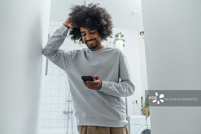 在家中浴室用智能手机的微笑男人，手放在头发上图片素材
