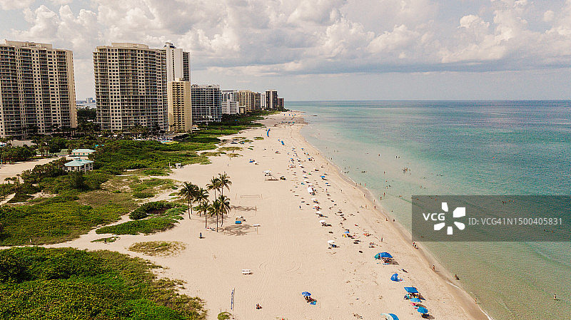 清澈的蓝绿色海浪航拍,人们在蓝绿色沙滩伞下放松,高大酒店和海滨房地产建筑位于佛罗里达州辛格岛海滩,2023年六月节中午图片素材