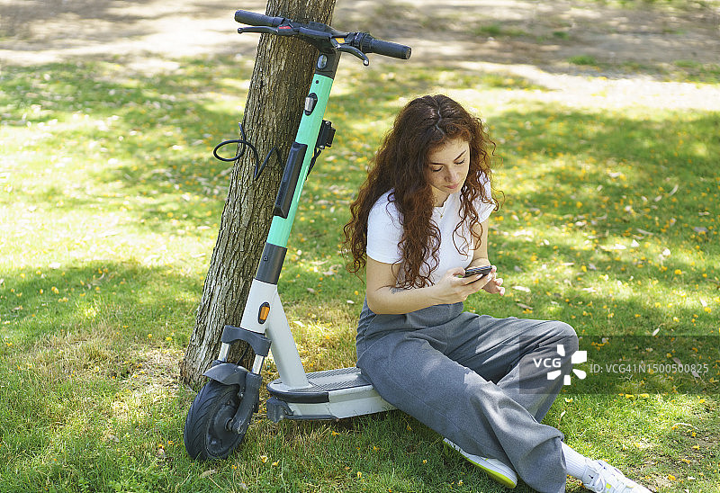 年轻女子将电动滑板车停在公园，坐在上面使用智能手机图片素材