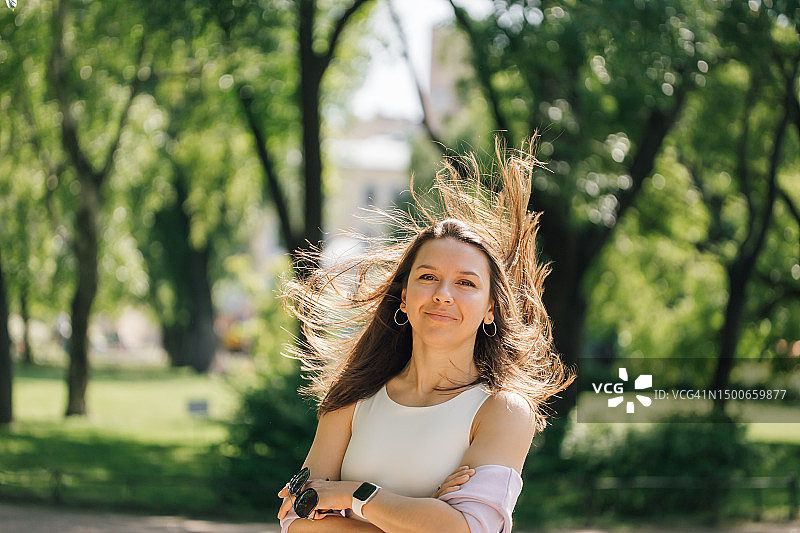 迷人的中年妇女，棕色长发被风吹乱，站在公园里看着镜头图片素材