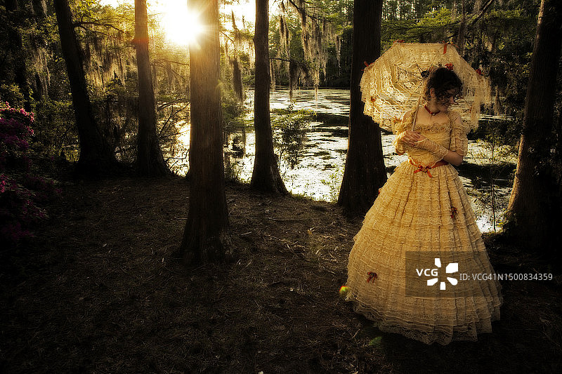 年轻女子身穿南方美女服装，包括大型华丽的时代礼服和阳伞，在湖边摆姿势图片素材