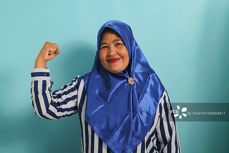 戴头巾的兴奋的亚洲妇女展示强壮的手势，弯曲手臂，自豪地微笑。蓝色背景图片素材
