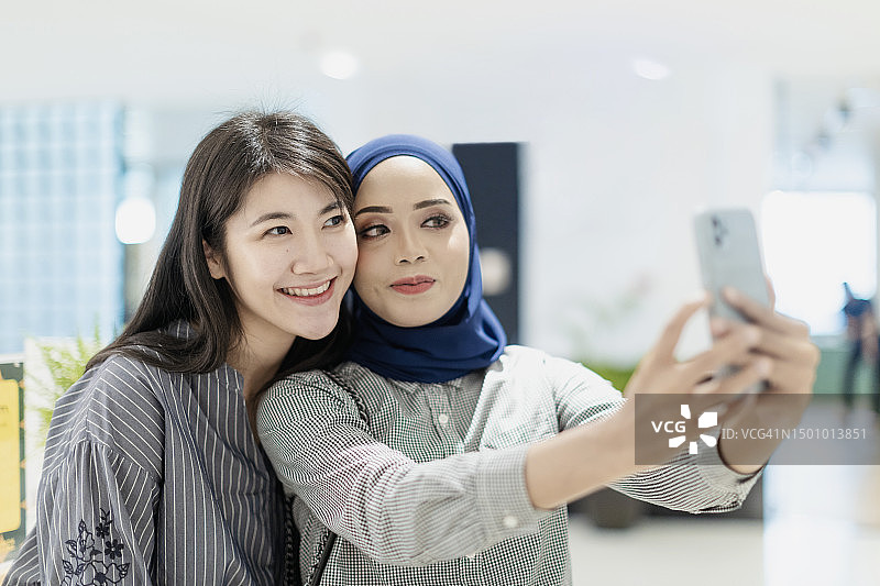 两名快乐的亚洲职业穆斯林女同事和中国女同事在咖啡休息期间使用智能手机在现代办公室自拍图片素材
