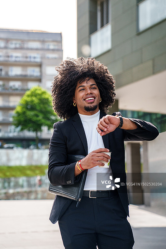 留着爆炸头、身穿优雅西装、拿着外带咖啡、在现代城市景观中看手表的拉丁裔商人图片素材