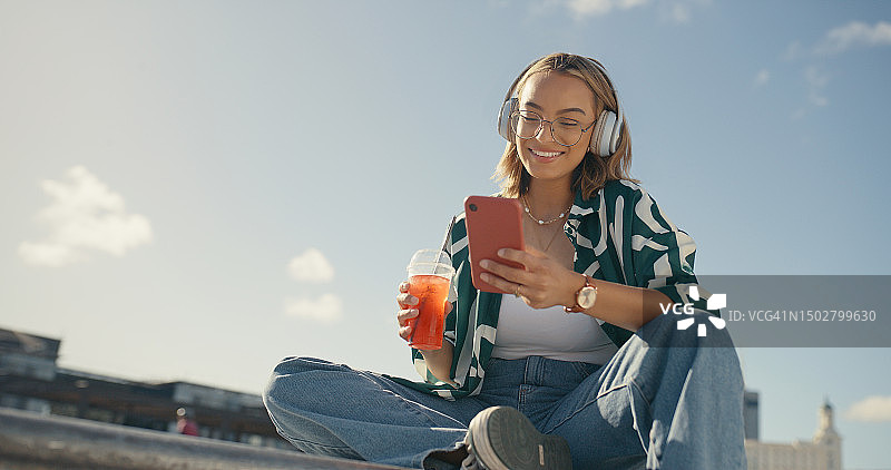 在城市里戴着耳机和手机，喝着冷饮的年轻女孩，在模拟天空中使用手机短信和社交媒体App。快乐的Z世代女性，城市网红，用智能手机在户外听音乐图片素材