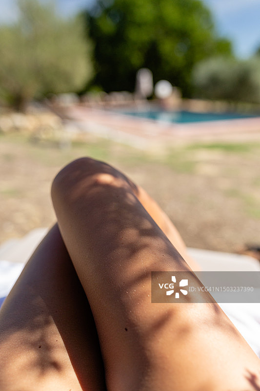 第一人称视角：女子在阳光明媚的泳池边享受日光浴，双腿出镜图片素材