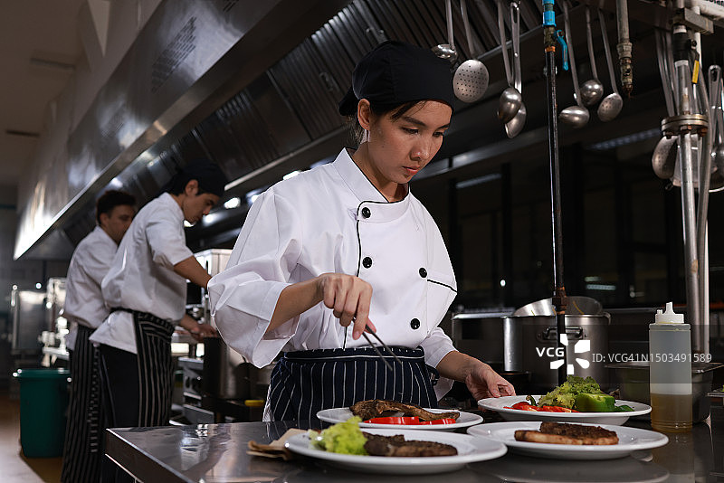 女厨师烹饪油炸肉类并摆放蔬菜装饰餐盘，酒店或餐厅厨房内的厨师团队，在炉子旁烹饪的多种族厨师团队图片素材